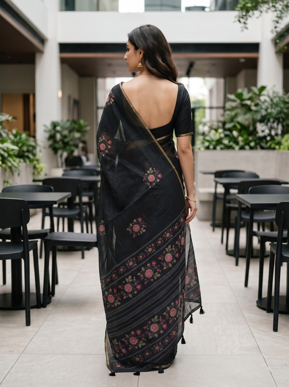 A woman stands in an outdoor patio setting wearing a black saree with floral prints and gold border details. The background features modern concrete structures, tables, and chairs.