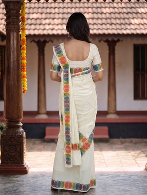 A woman viewed from behind wearing a white saree with colorful circular border prints, standing in an outdoor courtyard with carved pillars and a tiled roof in the background.