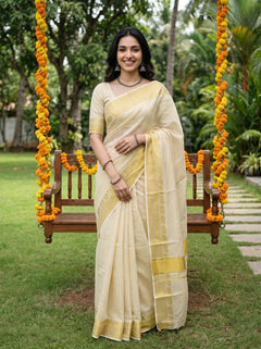 A woman wearing a cream-colored saree with yellow-gold border accents stands on a wooden swing decorated with orange marigold garlands in a lush green garden setting.