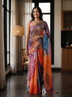 A woman wearing a colorful abstract-print saree in shades of orange, pink, purple, and blue, standing in a modern indoor room with large windows and curtains.