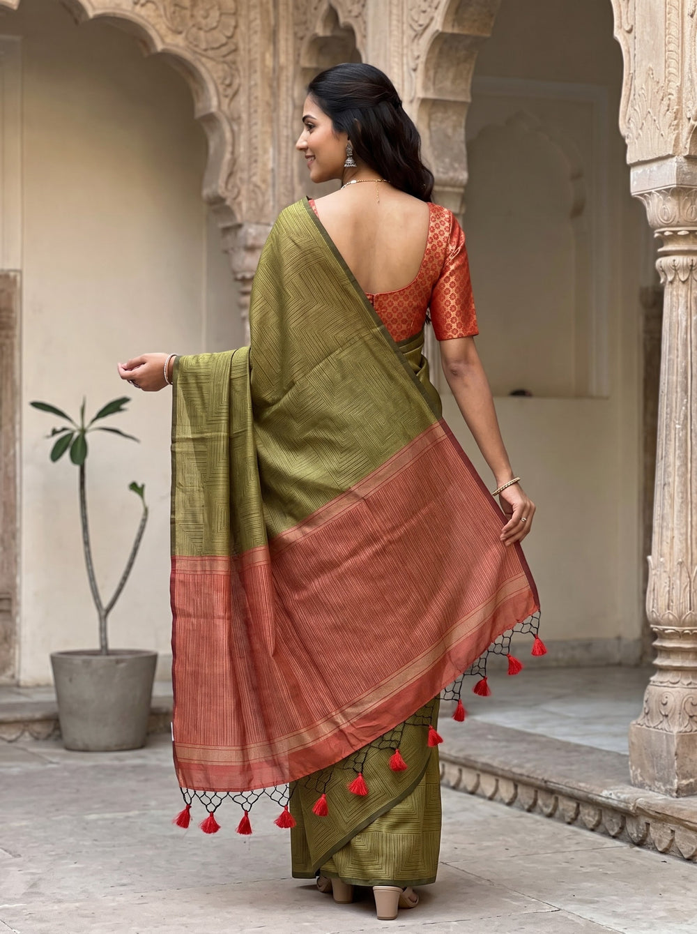 A woman modeling a sage green silk saree with a rust-orange border and matching blouse, standing in a traditional courtyard with arches.
