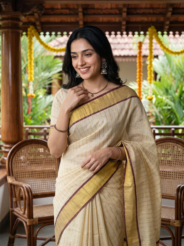 A woman wearing a beige and gold striped saree with brown borders, standing in a traditional porch setting with wooden chairs and greenery in the background.