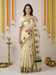 A woman modeling a cream-colored cotton saree with green and brown printed motifs and a striped border, standing in front of a neutral background decorated with hanging marigold garlands and brass lamps.