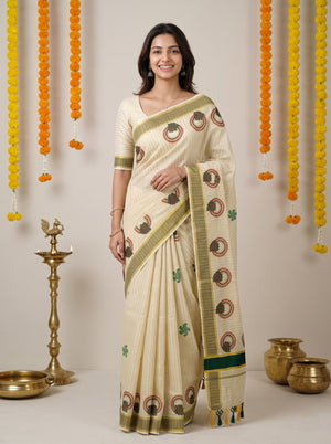A woman modeling a cream-colored cotton saree with green and brown printed motifs and a striped border, standing in front of a neutral background decorated with hanging marigold garlands and brass lamps.