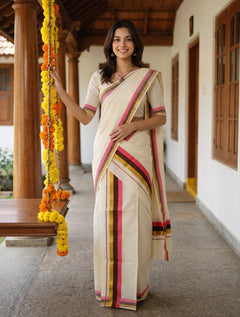 A woman wearing a cream-colored cotton saree with pink, yellow, and green striped borders stands on a traditional porch with wooden pillars and hanging marigold garlands.