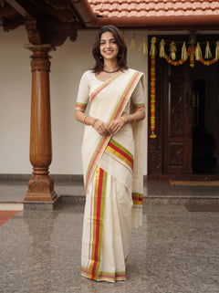 A woman wearing a cream-colored saree with red, yellow, and green stripes stands in front of a traditional house doorway with carved wooden pillars.