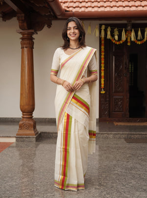 A woman wearing a cream-colored saree with red, yellow, and green stripes stands in front of a traditional house doorway with carved wooden pillars.