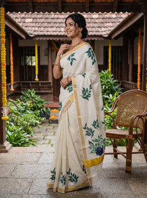 A woman wearing a cream-colored saree with green leaf patterns and gold borders, standing in a traditional courtyard with a wooden chair and tiled roof in the background.
