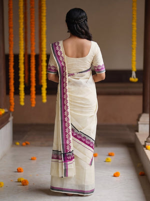 A woman viewed from behind wearing a cream-colored cotton saree with purple and black border patterns, standing in a corridor decorated with hanging marigold garlands.