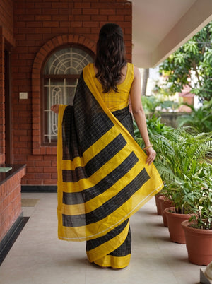 A woman viewed from behind wearing a yellow and black striped saree with a yellow sleeveless blouse, standing on a covered walkway beside potted plants.