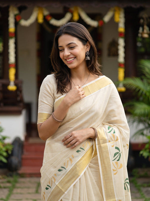 A woman wearing a cream-colored saree with yellow and green printed motifs, standing in front of a decorated traditional house.