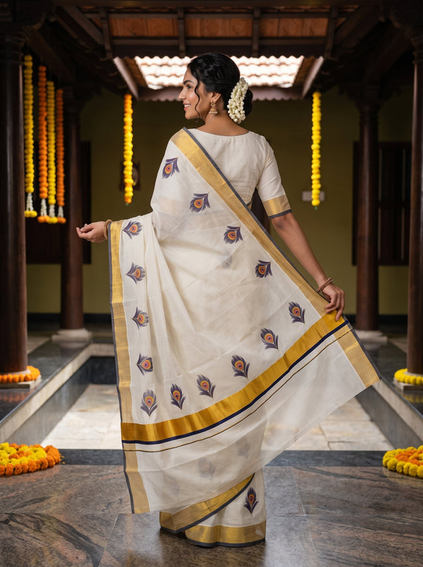A woman wearing a white cotton saree with gold borders and blue floral motifs, standing in a traditional courtyard.