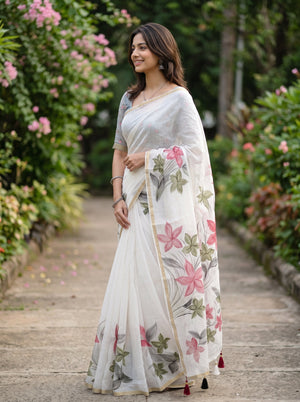A woman wearing a white saree with pink and green floral embroidery, standing on a stone path surrounded by lush green garden foliage and pink flowers.
