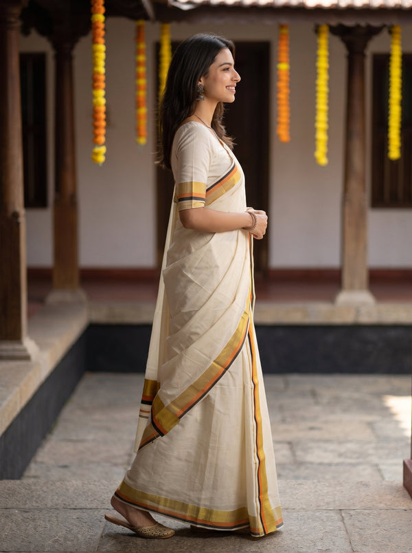 A woman wearing a cream-colored cotton saree with yellow, orange, and red border stripes, standing in profile in a traditional courtyard.