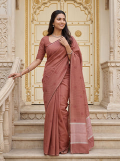 A woman wearing a dusty rose terracotta cotton saree with a matching printed blouse, standing on stone steps in front of an ornate cream-colored building door.