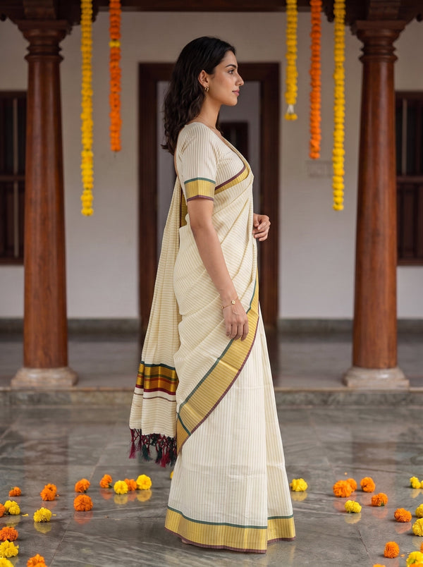 A woman wearing a cream-colored saree with yellow, green, and red border details stands in profile in a courtyard with wooden pillars and hanging marigold garlands.