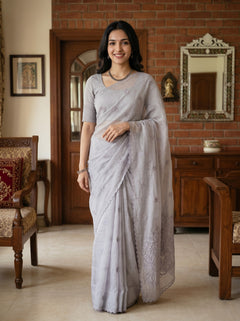 A woman wearing a light gray saree with a matching short-sleeve blouse, standing in a traditional indoor room with a brick wall, wooden furniture, and a mirror.