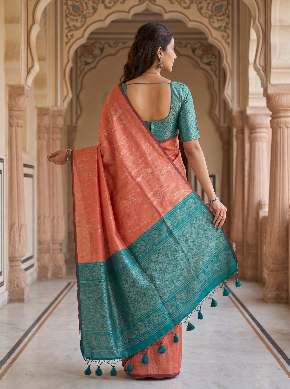 A woman stands in a stone arcade wearing a peach-orange saree with a teal green border and matching teal green short-sleeve blouse. The background features warm beige arches and columns.