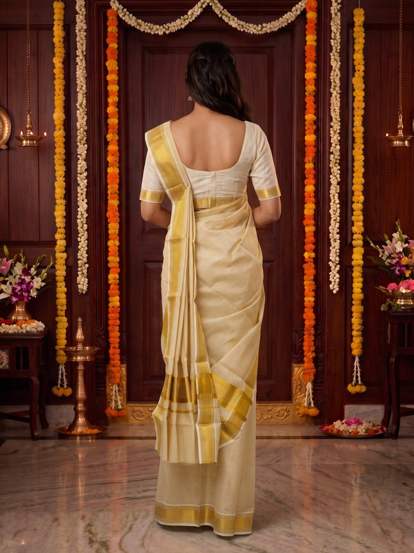 A woman viewed from behind wearing a cream and gold saree with a white blouse, standing in a decorated indoor setting with marigold garlands and a dark wooden door.