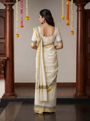 A woman viewed from behind wearing a cream-colored traditional saree with gold and black border patterns, standing in front of a wooden doorway decorated with hanging marigold garlands.