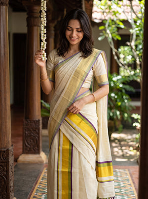 A woman wearing a cream-colored cotton saree with yellow, purple, and green stripes, standing in a traditional courtyard with wooden pillars and green foliage in the background.