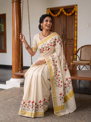 A woman wearing a cream-colored cotton saree with red floral prints and gold borders, sitting on a wooden swing in a traditional courtyard setting.