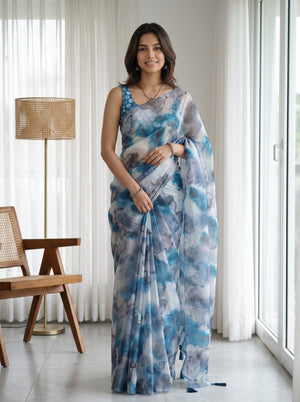 A woman modeling a blue and gray watercolor-patterned saree with a matching sleeveless blouse in a bright, modern room with white curtains and a wooden chair.