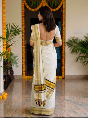 A woman viewed from behind wearing a white saree with yellow and black border stripes, standing in an indoor doorway decorated with yellow marigold garlands.