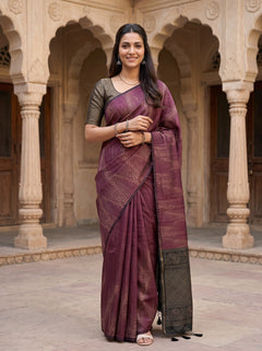 A woman wearing a purple and gold patterned saree with a dark grey patterned blouse stands in front of a historic stone building with arches.