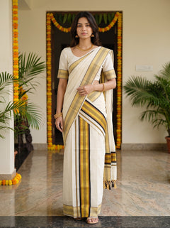 A woman modeling a traditional Indian saree in an indoor setting with marble flooring and potted plants. The saree is off-white/cream with prominent vertical stripes in black, mustard yellow, and brown, paired with a matching cream blouse.