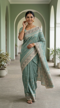 Woman in a light blue saree with a white blouse standing in a hallway with arches and plants.