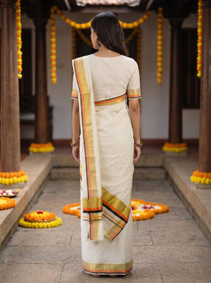 A woman viewed from behind wearing a cream-colored cotton saree with a gold, orange, and red striped border, standing in a traditional courtyard decorated with marigold garlands.