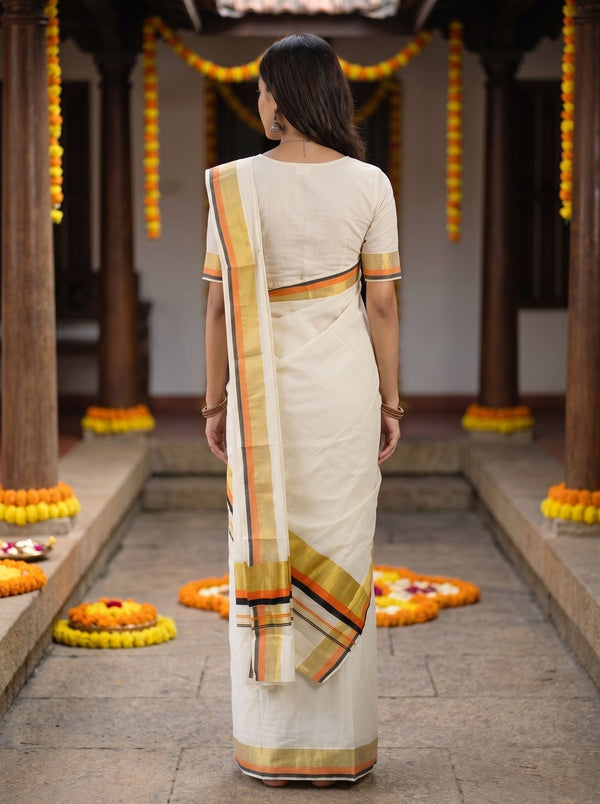 A woman viewed from behind wearing a cream-colored cotton saree with a gold, orange, and red striped border, standing in a traditional courtyard decorated with marigold garlands.