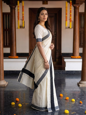 A woman wearing a cream-colored cotton saree with dark gray borders and a matching short-sleeve blouse, standing in a traditional courtyard with wooden pillars and marigold garlands.