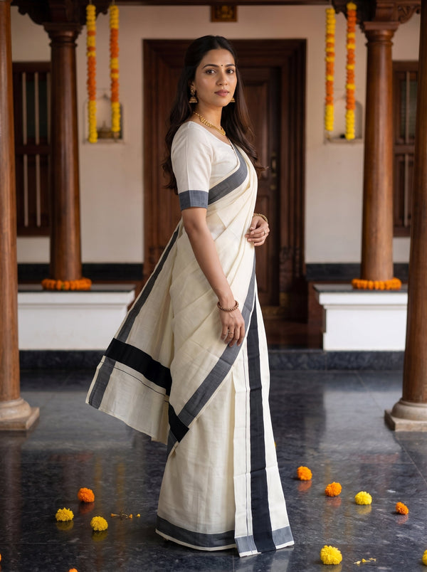 A woman wearing a cream-colored cotton saree with dark gray borders and a matching short-sleeve blouse, standing in a traditional courtyard with wooden pillars and marigold garlands.