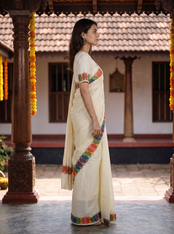 A woman wearing a cream-colored traditional saree with colorful floral border prints, standing in profile under a wooden pavilion.