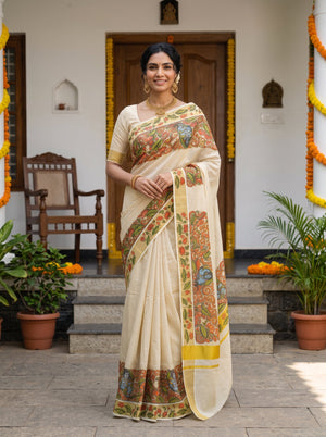 A woman stands outdoors in front of a doorway wearing a cream-colored saree with a colorful floral border and a matching printed blouse. The setting includes potted plants, stone steps, and yellow garlands.