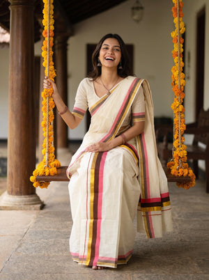 A woman wearing a cream-colored cotton saree with pink, yellow, and green stripes sits on a wooden swing decorated with marigold garlands in a traditional courtyard.