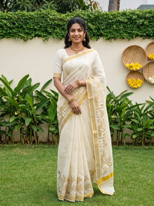 A woman stands outdoors on a lawn wearing a cream-colored saree with gold floral patterns and a matching cream blouse. The background features green plants, a beige wall, and decorative woven baskets with yellow flowers.