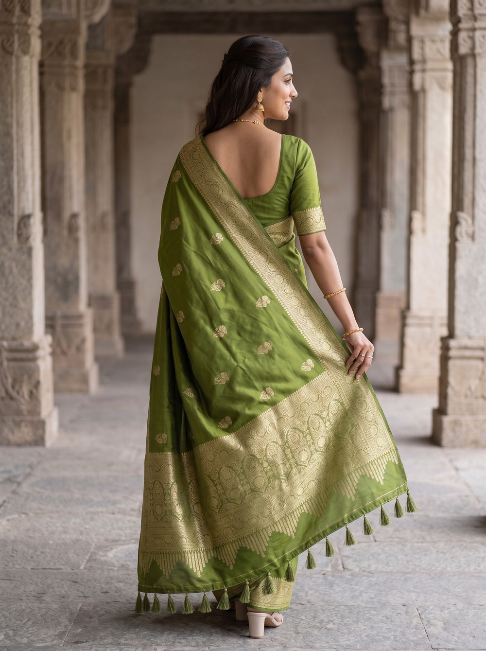 A woman wearing a green Banarasi silk saree with gold zari floral motifs and a wide gold border, standing in a stone corridor.