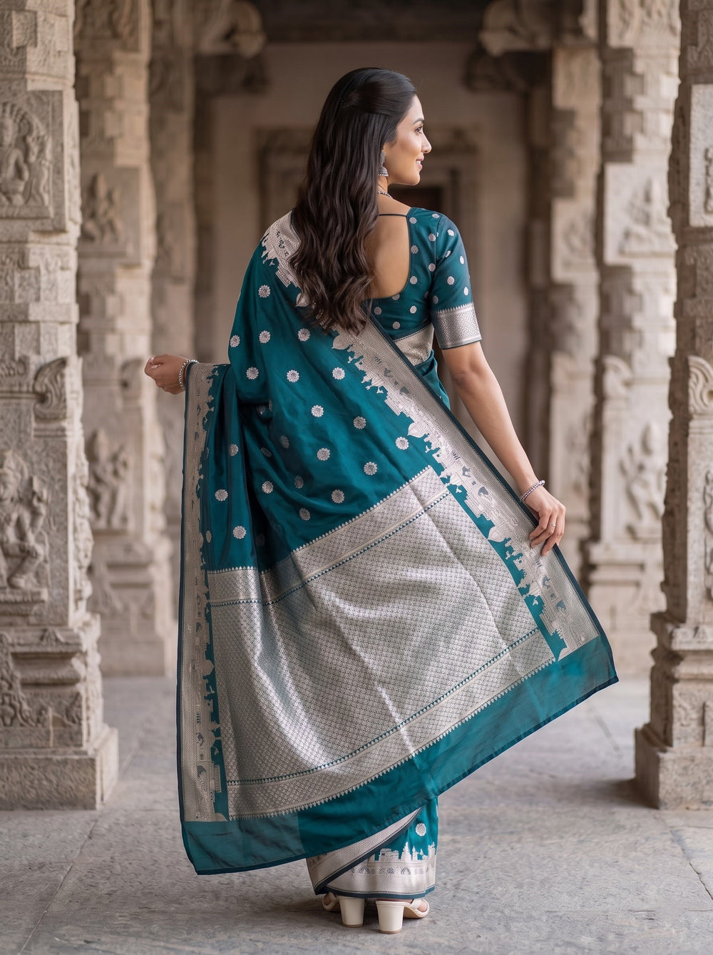 A woman wearing a teal blue and silver saree stands in a stone corridor with carved pillars. The saree has silver polka dots and a wide silver border with a white and silver patterned drape.