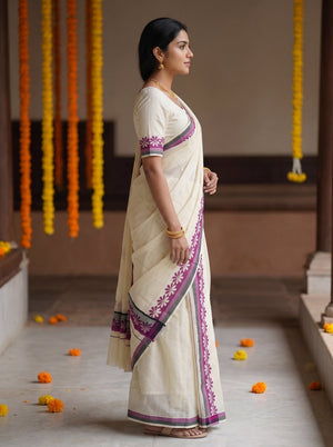 A woman wearing a cream-colored cotton saree with purple and green border embroidery, standing in a traditional courtyard decorated with orange marigold garlands.