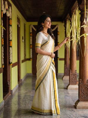 A woman wearing a cream-colored cotton saree with yellow and green border stripes stands in a traditional corridor with wooden pillars and hanging green leaves.