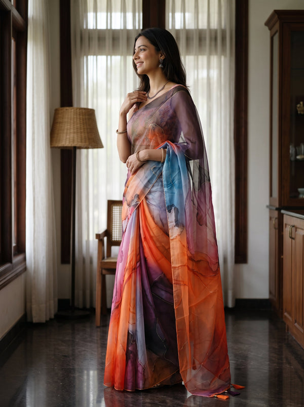 A woman wearing a flowing multicolor printed saree stands indoors near large windows with sheer curtains. The saree has an abstract watercolor-like pattern in orange, blue, purple, pink, and white, with a sheer blouse and draped pallu.