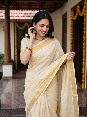 A woman wearing a light beige and yellow saree stands in a traditional courtyard, holding out the pallu to display its border pattern.