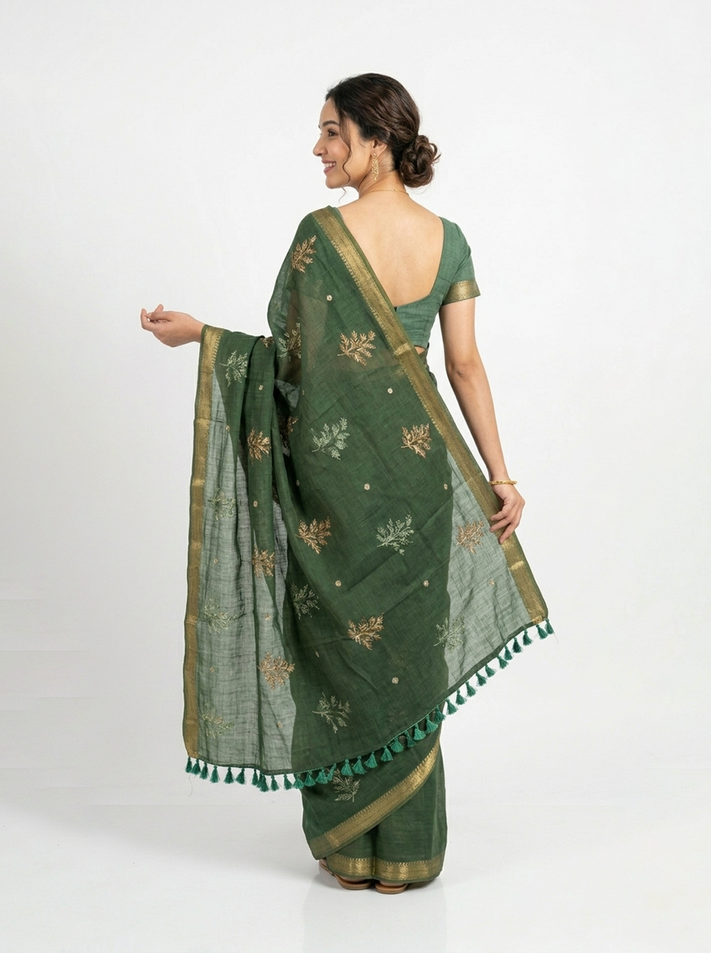Woman in a green saree standing in a room with a lamp and books.