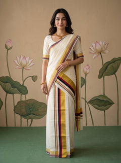 A woman modeling a cream-colored cotton saree with yellow, maroon, and gold stripes, standing in front of a beige wall decorated with lotus motifs.
