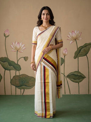 A woman modeling a cream-colored cotton saree with yellow, maroon, and gold stripes, standing in front of a beige wall decorated with lotus motifs.