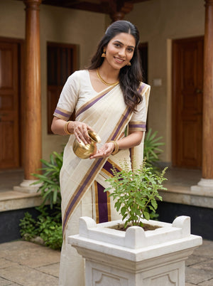 A woman wearing a cream-colored traditional saree with purple and gold borders stands in a courtyard, holding a small brass pot over her hand next to a white planter with a green plant.