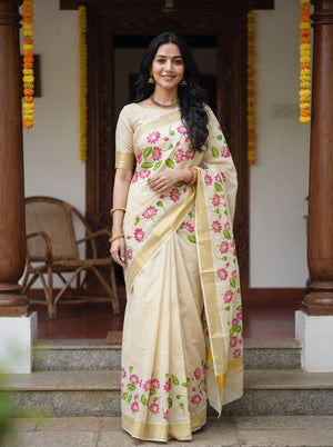 A woman wearing a cream-colored saree with pink floral prints and green leaves, standing in front of a traditional house doorway decorated with yellow marigold garlands.